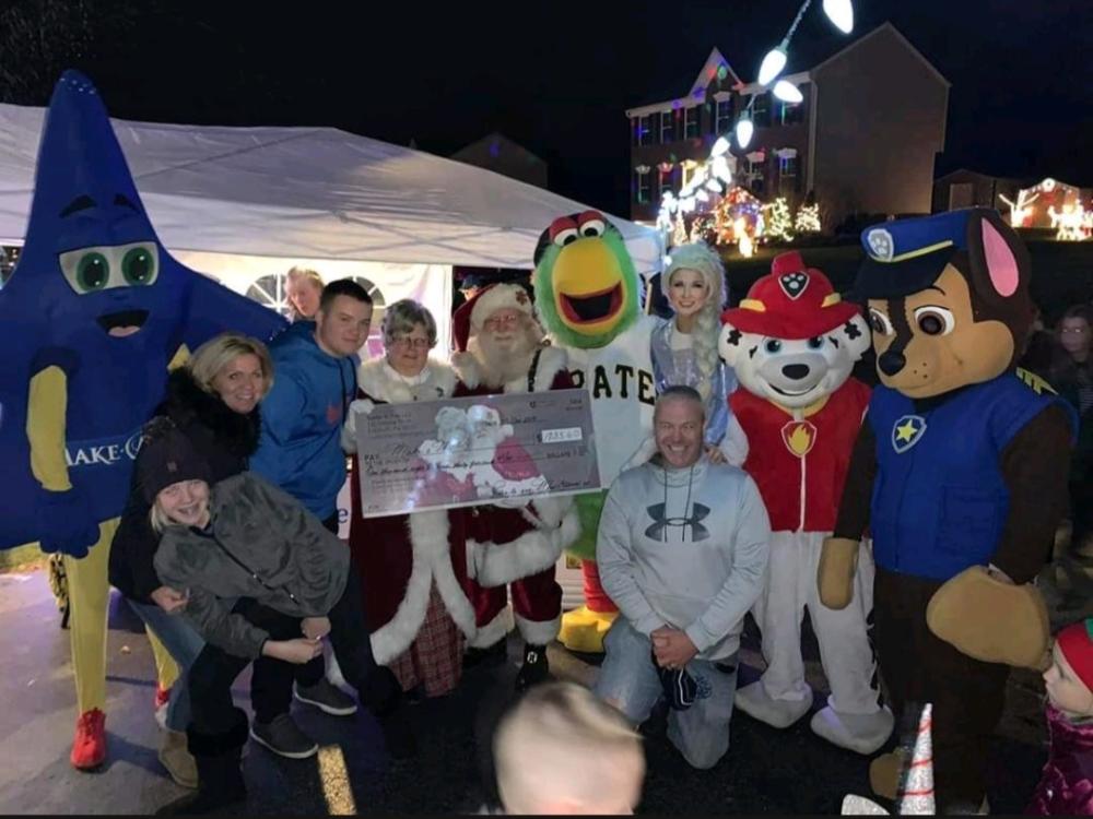 A group of people pose together at a holiday event with Santa and Mrs. Claus, several costumed characters, and a large ceremonial check, with Christmas lights decorating the background.