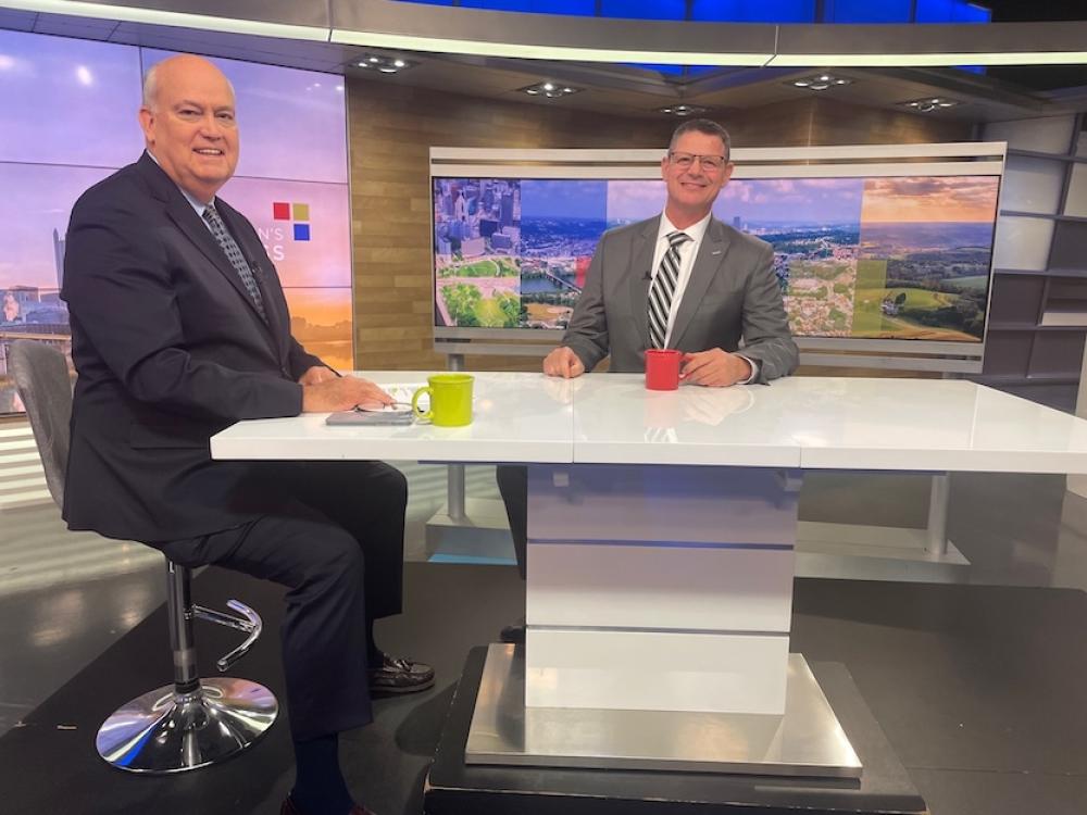Peoples President Mike Huwar with Bill Flanaganin at a news studio desk, smiling toward the camera, with colorful cityscape images displayed on a screen behind them.