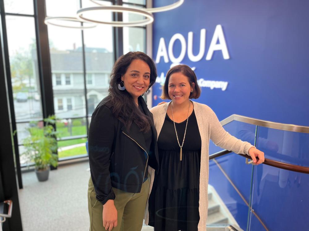 Sarah (left) and Blair (right) standing together and smiling inside an Aqua office building, with large windows and a blue wall in the background.