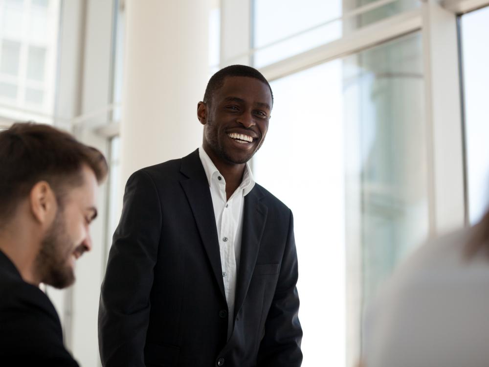 A man in a suit smiles while standing in a bright, modern office with large windows.