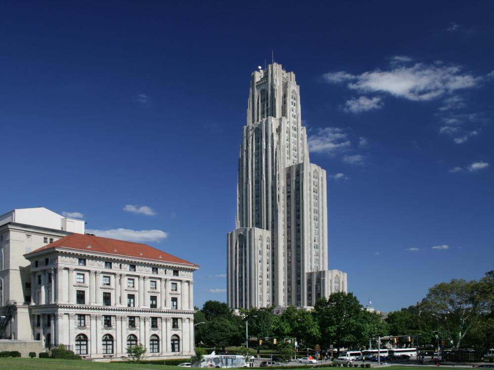 The Cathedral of Learning rises above surrounding buildings and trees under a clear blue sky.