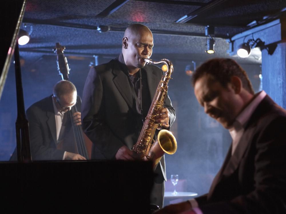 Three musicians perform in a dimly lit jazz club, with a saxophonist at center, a pianist in the foreground, and a bassist in the background.