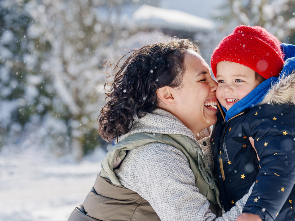 A woman and a young child bundled in winter clothing smile and hug outdoors as snow falls around them.