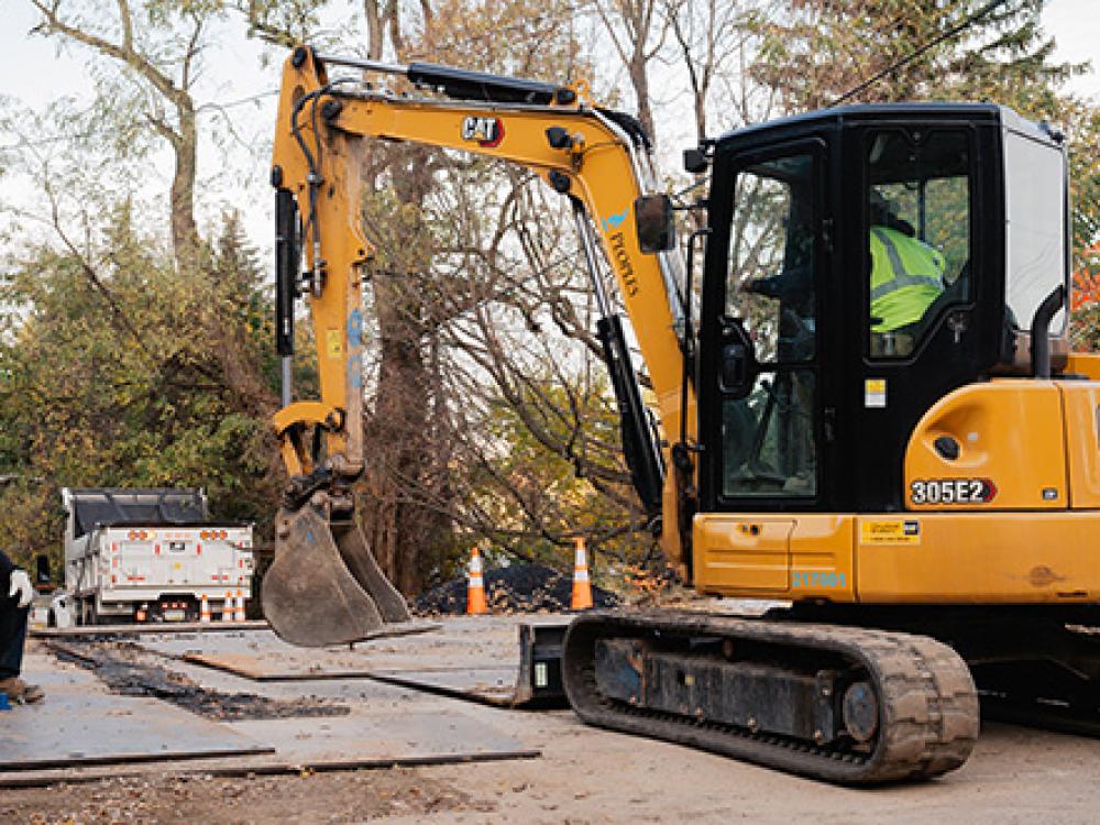 A construction worker kneels near a small excavator as it digs on a street.