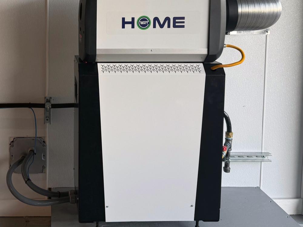 A standalone home energy unit labeled “WATT HOME” is mounted on a platform against a wall, with ventilation ducting attached on the right and connections running to the unit.