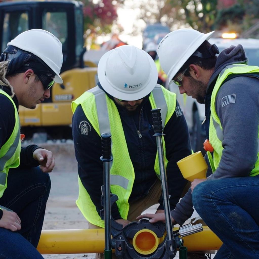 Three workers in hard hats and safety vests kneel around a yellow pipeline, installing or inspecting pipe connections at a work site.