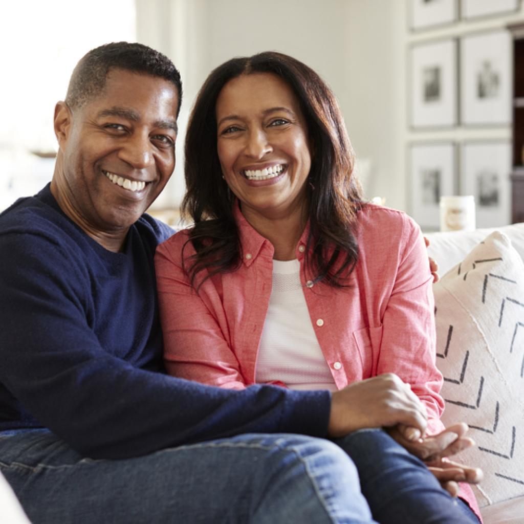 A smiling couple sits closely on a beige sofa in a bright living room, with framed photos and a cabinet in the background.