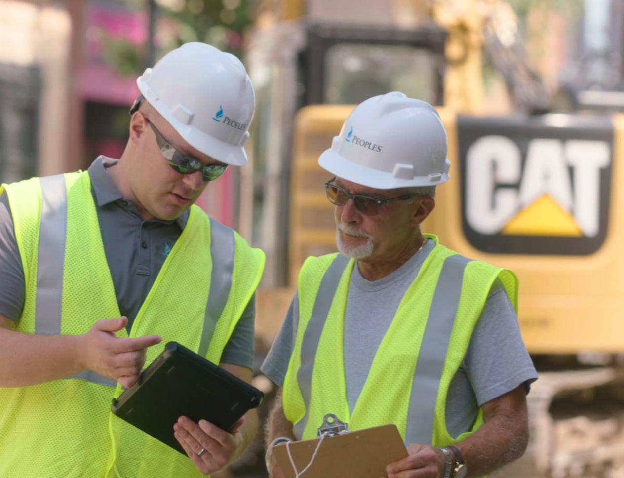 Two construction workers wearing white hard hats and high-visibility vests review information on a tablet at a city street work site.