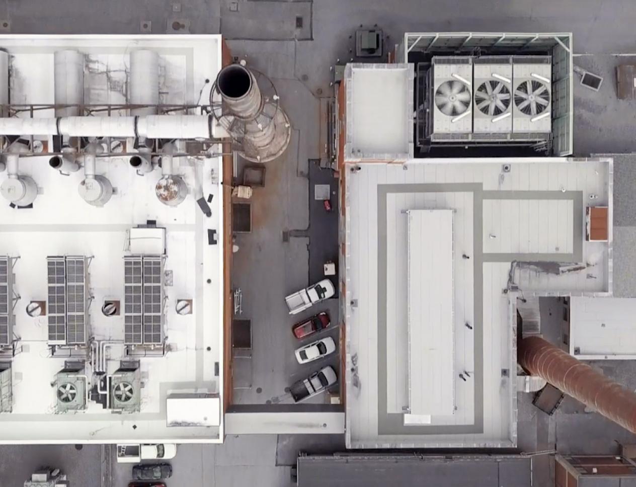 Aerial view of an industrial facility rooftop showing large ventilation units, pipes, exhaust stacks, and adjacent buildings with parking areas.