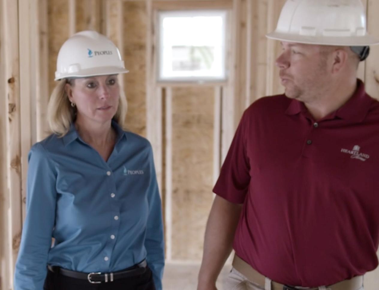 Two people wearing hard hats walk through the interior of an unfinished house, discussing construction work.