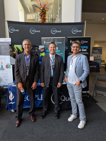 Three men standing and smiling in front of an HQuest booth at an indoor event. They are dressed in business attire and wearing conference badges, with informational banners in the background.