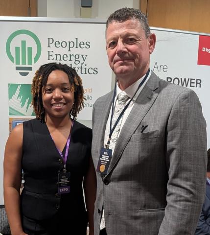 Two people standing and smiling in front of a Peoples Energy Analytics display at an event