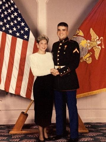 Vintage photo of a U.S. Marine in dress uniform standing with a woman in front of American and Marine Corps flags.