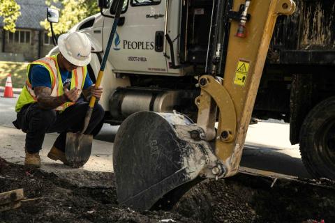 Utility worker in a hard hat and safety vest kneels beside a trench, holding a shovel next to excavation equipment on a residential street.