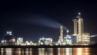 Illuminated industrial facility at night with towers, tanks, and smokestacks beside a reflective body of water.