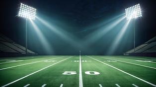 Empty football field at night illuminated by bright stadium lights, with the 50-yard line centered and empty bleachers in the background.