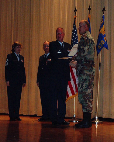 Service members in uniform stand on a stage as one presents an award or certificate to Brent Reech, with U.S. flags displayed behind them.